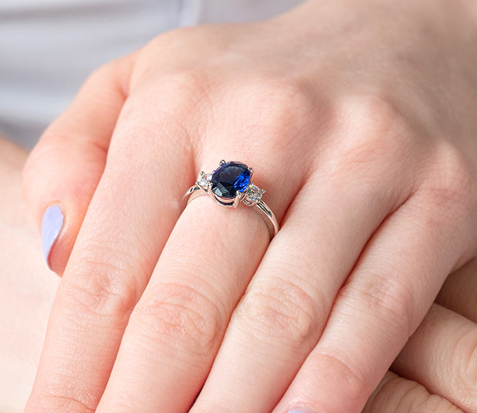 Oval cut blue ring with clear side stones in rhodi